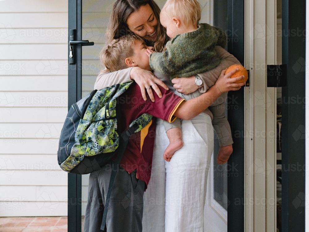 Aussie mum carrying toddler boy while hugging older son leaving for school at the porch - Australian Stock Image