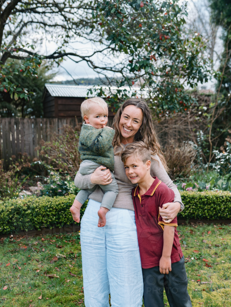 Aussie mum stands in the garden carrying toddler while hugging the older son - Australian Stock Image