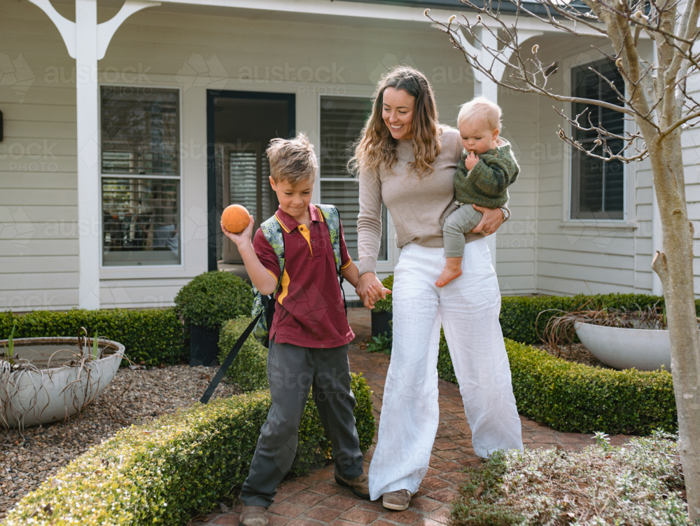 Aussie mum walking with young son while carrying her toddler on her hip to do school drop-off - Australian Stock Image