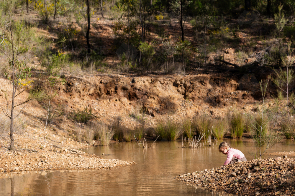 Aussie toddler playing by edge of waterhole in remote Australian bushland in afternoon sunlight - Australian Stock Image
