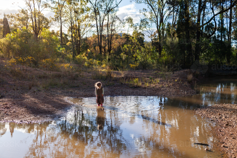 Aussie toddler playing by edge of waterhole in remote Australian bushland in afternoon sunlight - Australian Stock Image