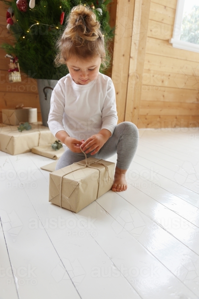 Little girl opening present with Christmas tree in background - Australian Stock Image