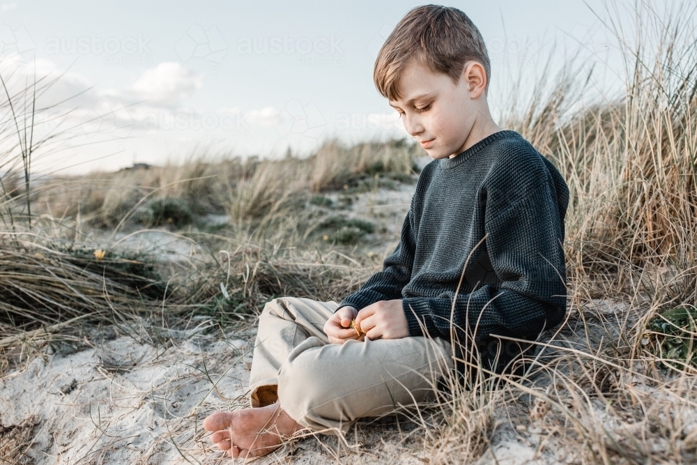 autistic boy sitting on a sand dune looking at his hands which are holding a yellow flower - Australian Stock Image