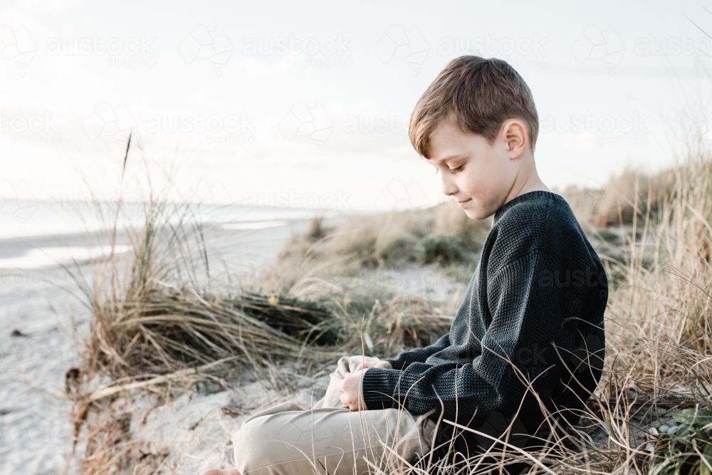 Autistic boy sitting on the grass on sand dunes at the beach - Australian Stock Image