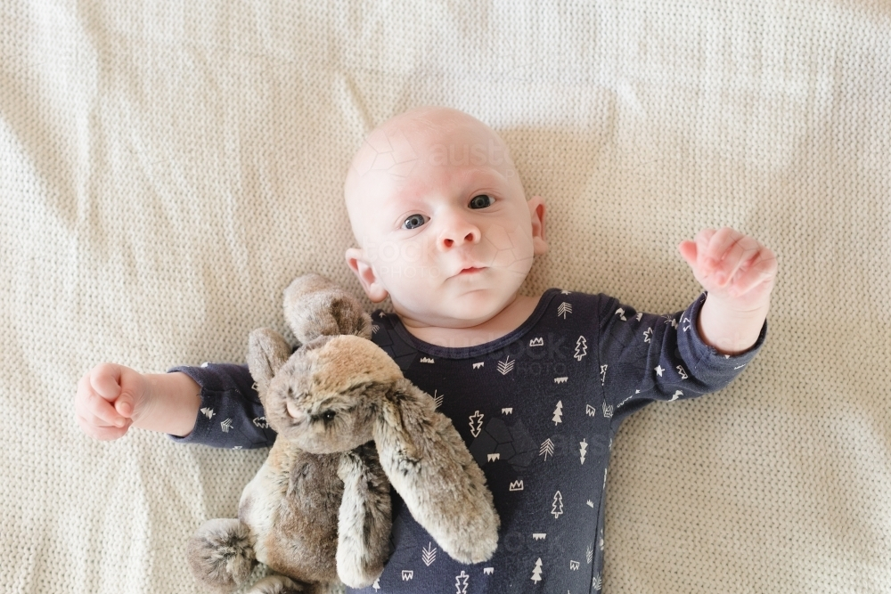 Baby boy with soft toy rabbit lying on bed - Australian Stock Image