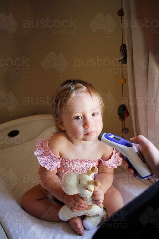 Baby girl holding a toy giraffe while having her temperature taken - Australian Stock Image