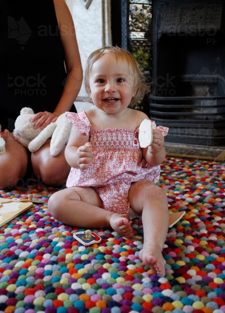 Baby girl sitting on a rug, playing with toys and smiling at camera. - Australian Stock Image