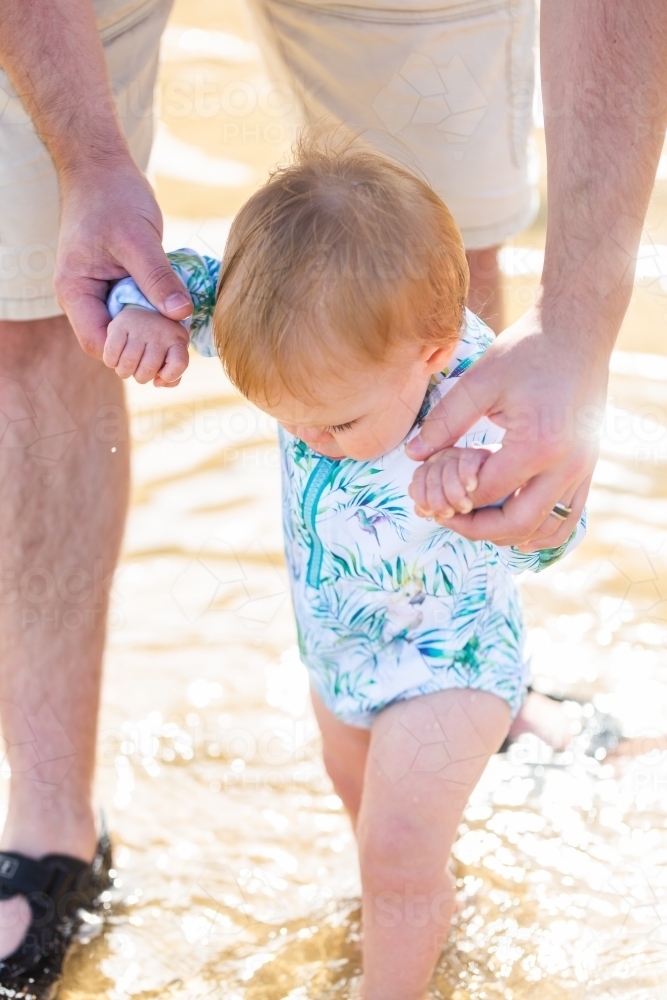 Baby holding onto fathers hands walking through sparkling ocean water at the seaside - Australian Stock Image