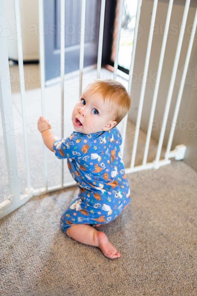 Baby looking over shoulder kneeling at child proof fence gate - Australian Stock Image