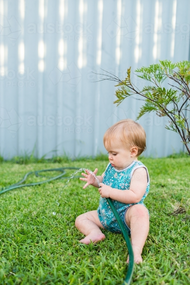 Baby on green backyard lawn playing with water in summer - Australian Stock Image