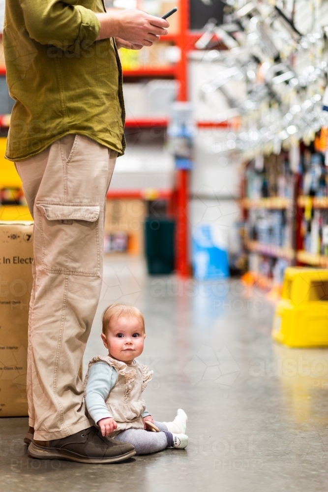 Baby sitting at fathers feet in hardware store - Australian Stock Image