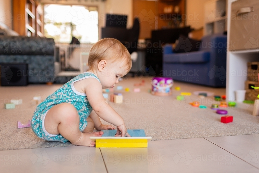 Baby sitting on floor pointing to page in book she is reading among mess of toys - Australian Stock Image