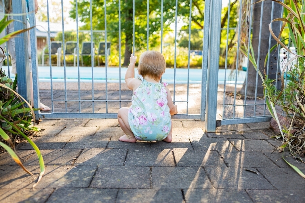 Baby squatting at pool gate on summer afternoon - Australian Stock Image