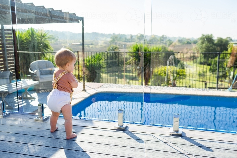 Baby standing at glass pool fence looking at water safe from drowning - Australian Stock Image