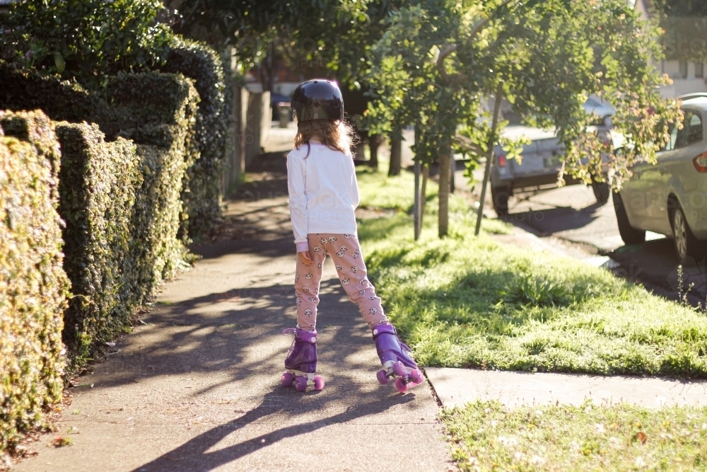 Back shot of a girl with helmet while roller skating on the side walks with trees, grass and hedge - Australian Stock Image
