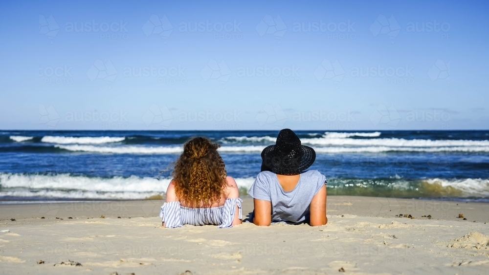 Image of Back shot of two girls sitting on beach - Austockphoto