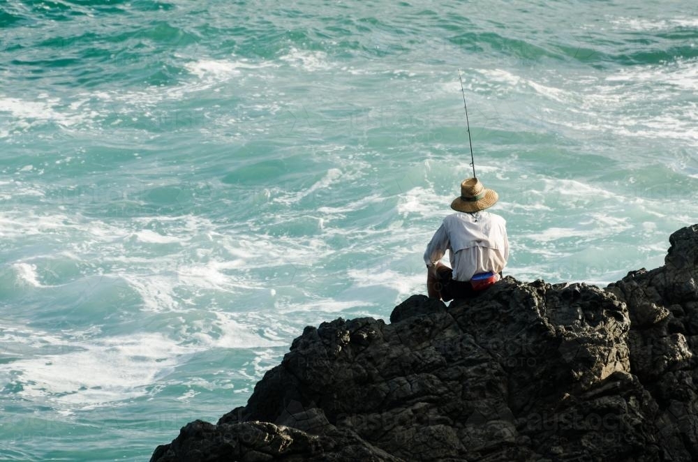 Back view of man fishing in the ocean from rocks - Australian Stock Image
