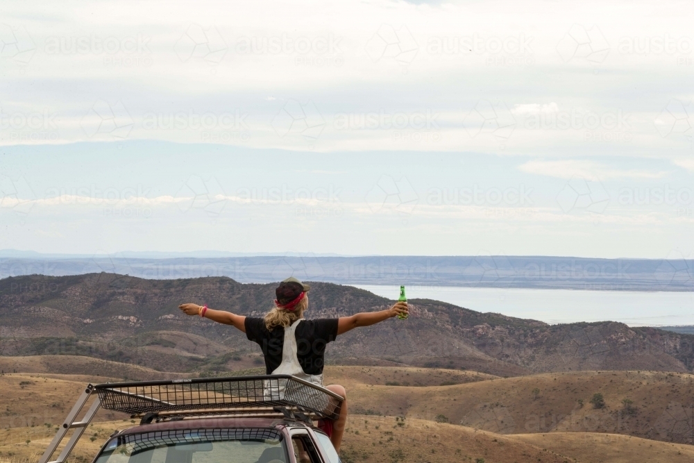 Back view shot of a woman holding a green bottle sitting on top of the car looking at the mountain - Australian Stock Image