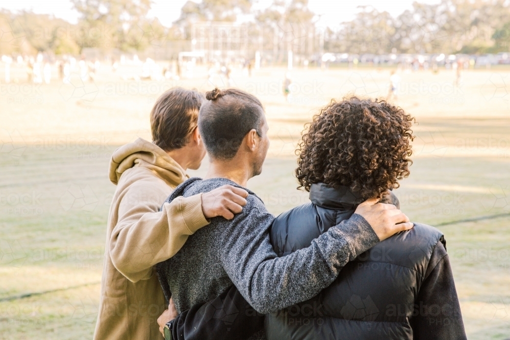 back view shot of three men one curly hair and one with a manbun with arms over each other's back - Australian Stock Image