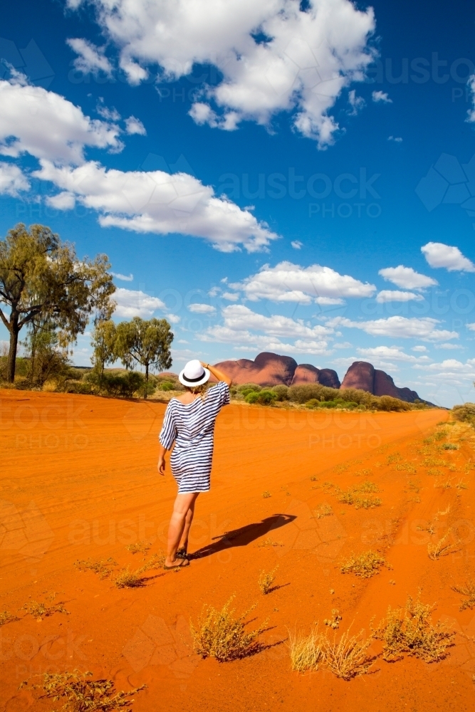 back view shot of woman walking on dirt road wearing striped dress and a hat with sunny clear skies - Australian Stock Image