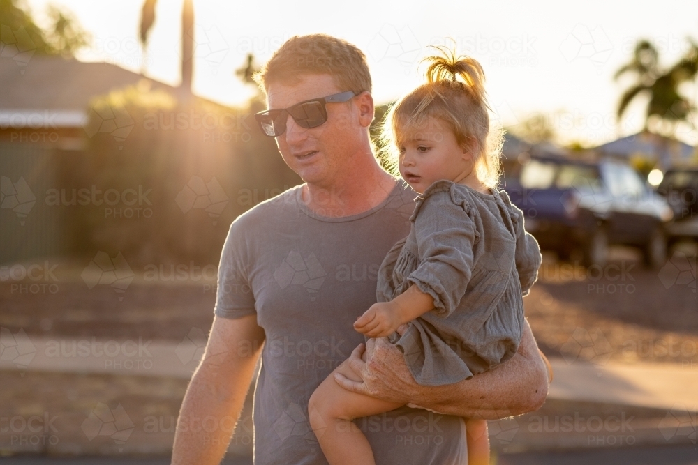 backlit father wearing sunnies carrying toddler on hip - Australian Stock Image