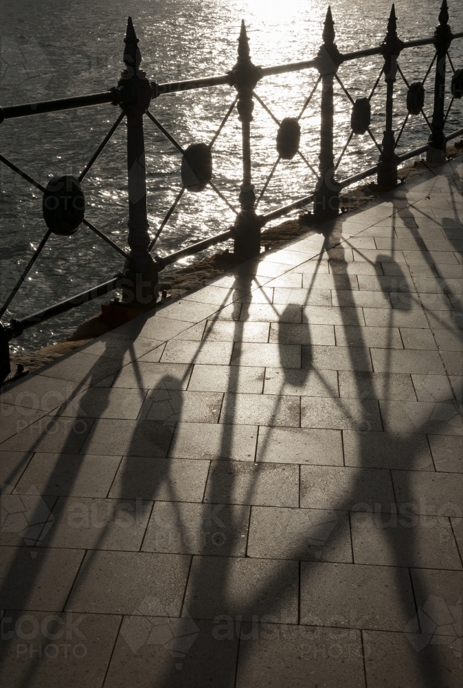 Backlit heritage safety fence throwing geometric shadows on paving with sunlit water behind - Australian Stock Image