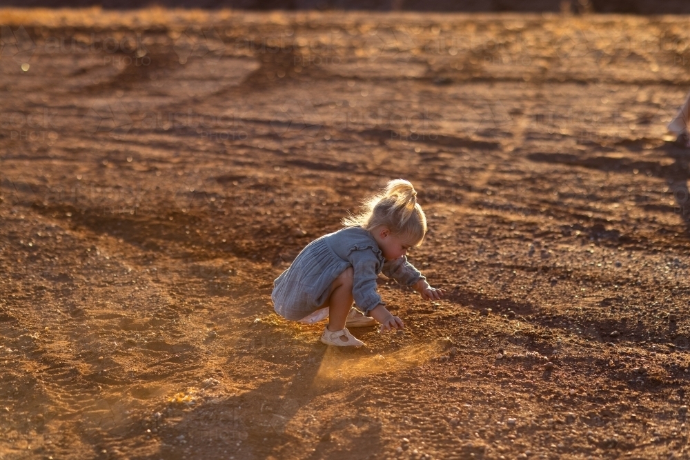 backlit little kid squatting down playing with stones on the ground - Australian Stock Image