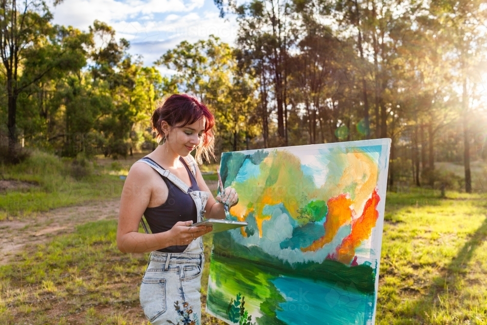 Backlit young artist in forest clearing working on large landscape painting - Australian Stock Image