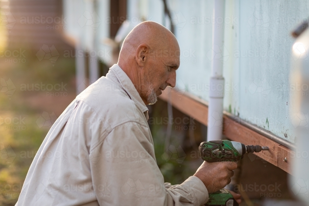 bald man using cordless drill for maintenance on dilapidated house - Australian Stock Image