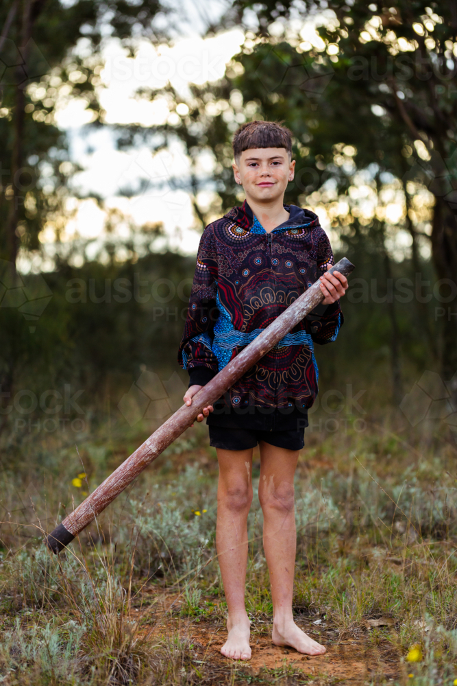 barefoot aboriginal boy holding didgeridoo instrument in native bushland with bokeh background - Australian Stock Image