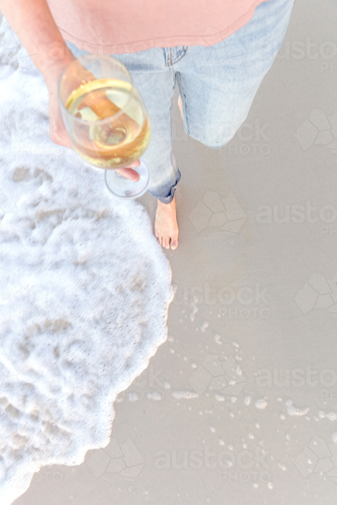 barefoot woman walking along beach holding glass of wine - Australian Stock Image