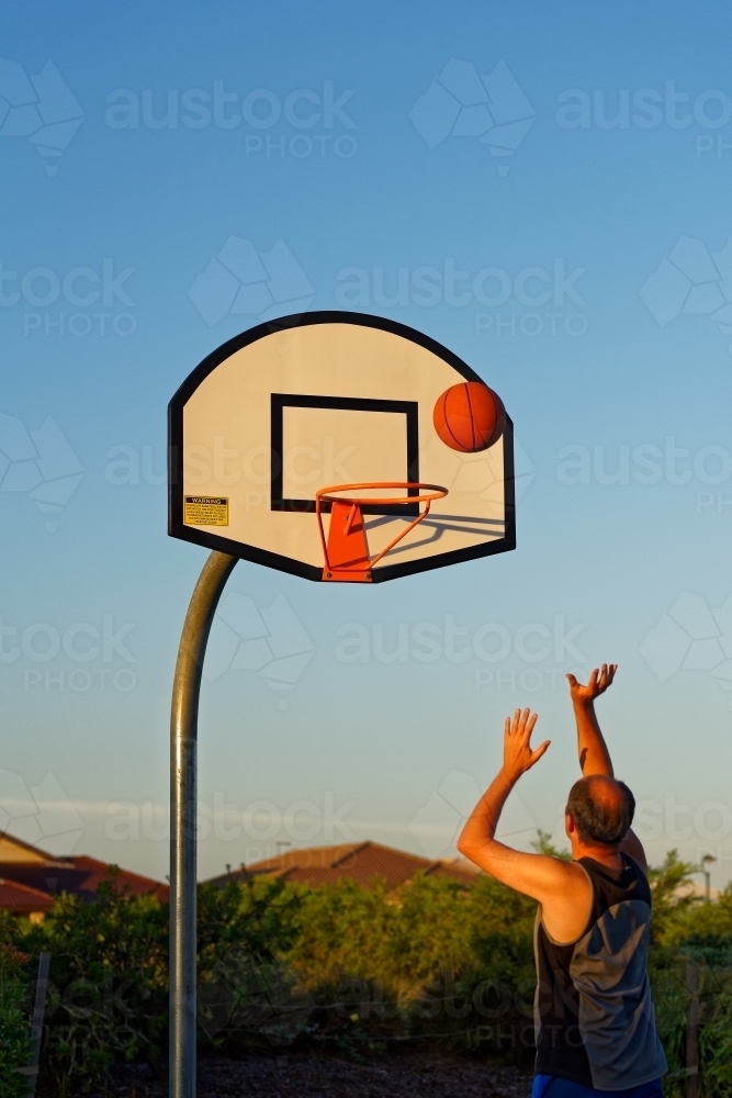 Basketball player shooting basket at sunset in outdoor park - Australian Stock Image