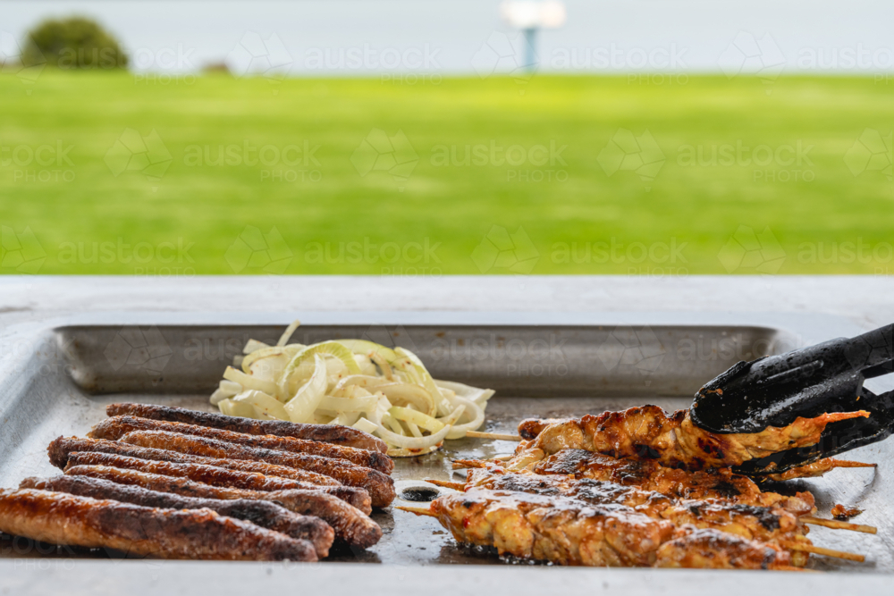 BBQ tray with grilled chicken, sausages, and caramelized onions cooking on it in park - Australian Stock Image