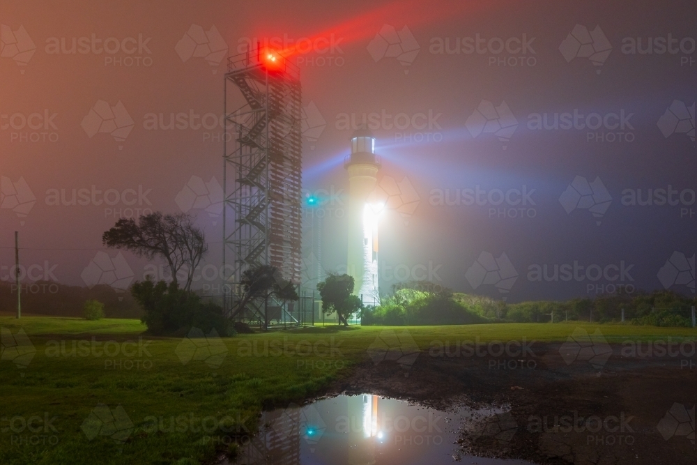 Beams of light from a lighthouse and beacon tower reflected in a pool of water on a dark foggy night - Australian Stock Image