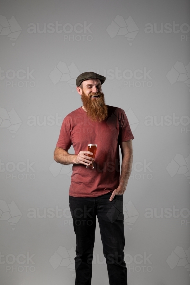 Bearded man in tweed flat cap standing holding a beer. - Australian Stock Image