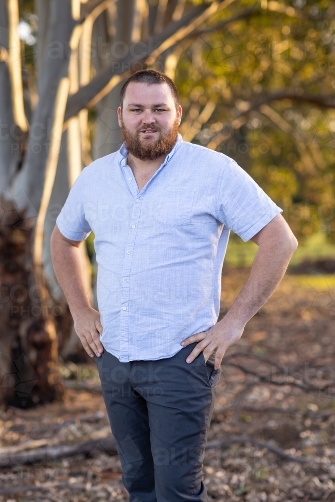 bearded man with hands on hips standing outdoors with trees in background - Australian Stock Image