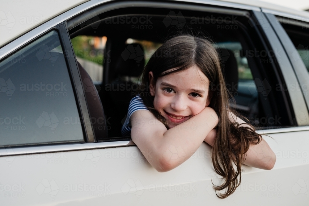 Beautiful, happy young girl leaning out the open window of the family car parked in the driveway - Australian Stock Image