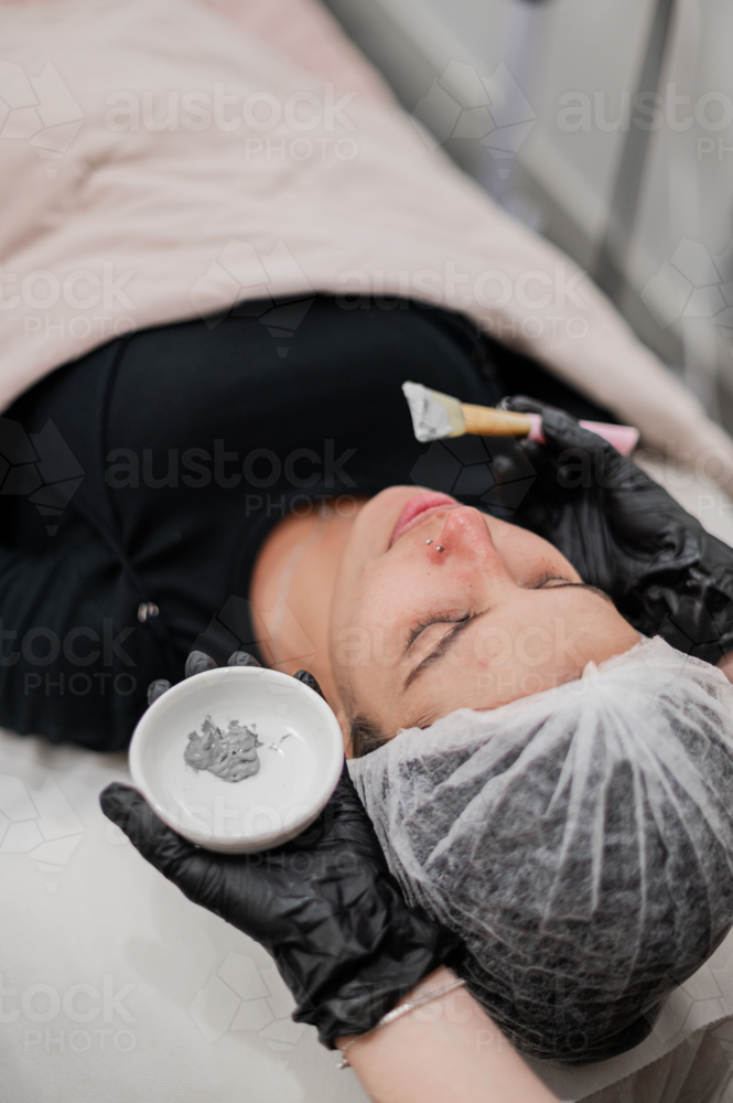 Beauty therapist applying a grey facial mask to a client lying on a treatment bed - Australian Stock Image