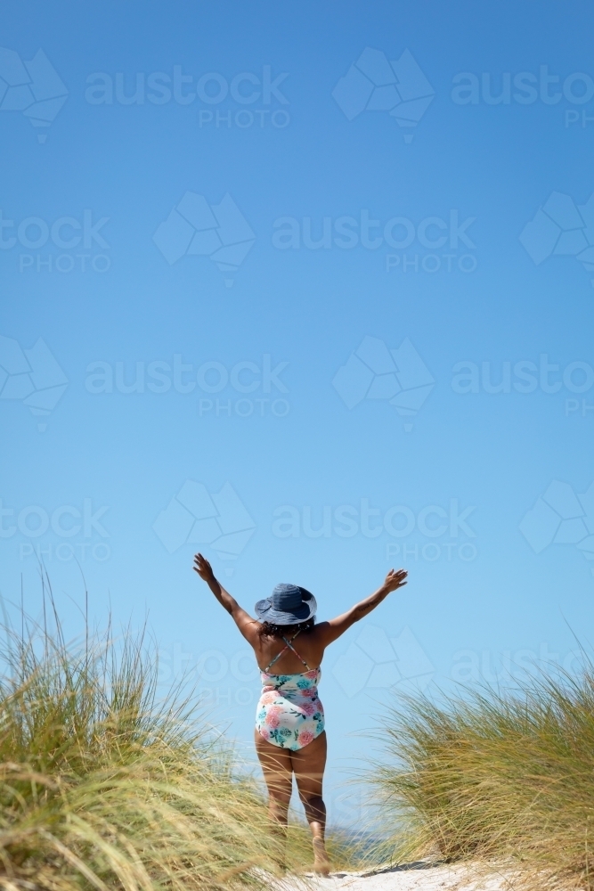 Big blue sky and woman in swimming costume walking towards the beach with arms raised - Australian Stock Image
