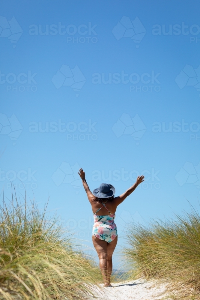 Big blue sky and woman in swimming costume walking towards the beach with arms raised - Australian Stock Image