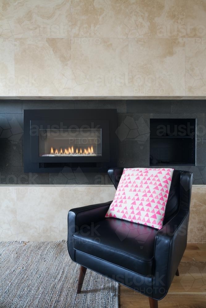 Black leather armchair and pink cushion next to warm cosy fireplace - Australian Stock Image