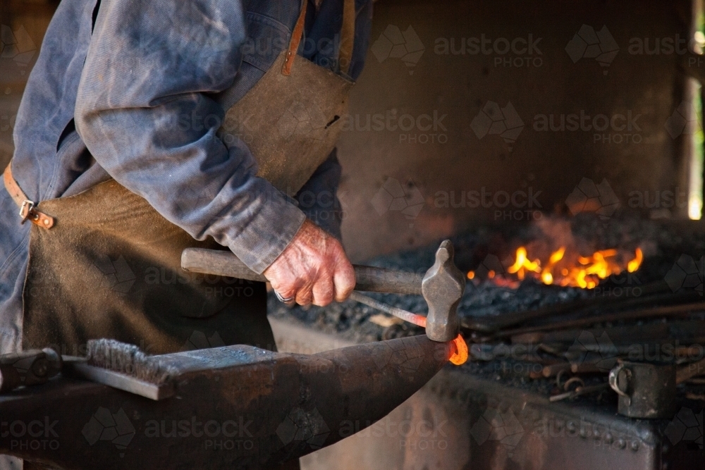 Blacksmith working hot metal with a hammer and anvil beside beside forge fire - Australian Stock Image