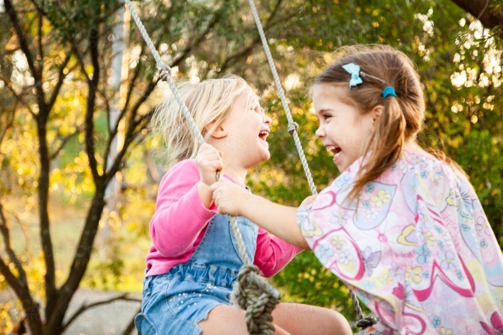 Blond and brunette little girls laughing on swing together - Australian Stock Image