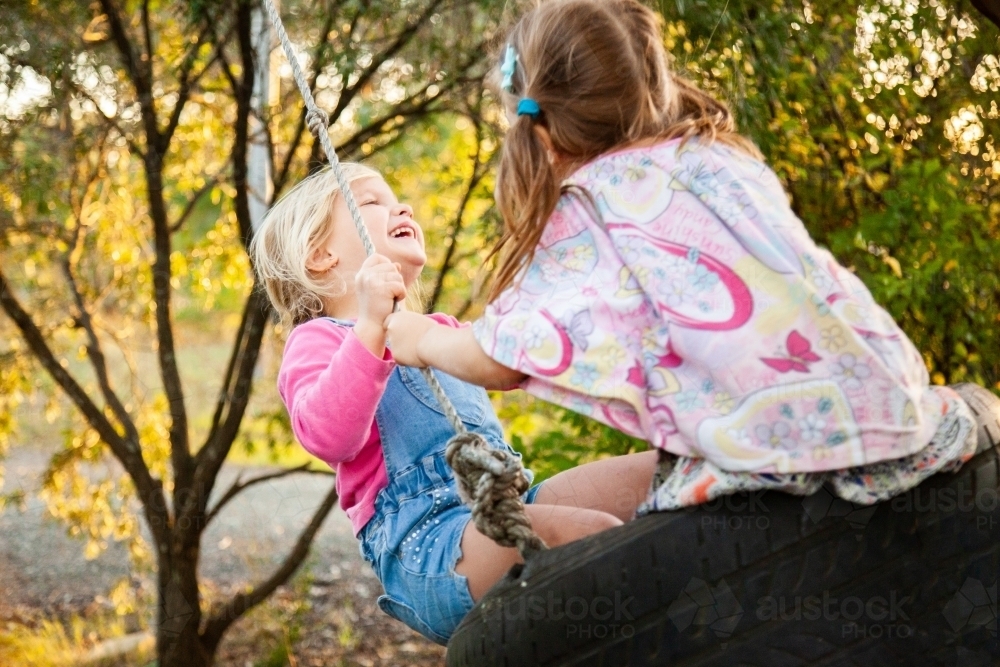 Blond and brunette little girls laughing on swing together - Australian Stock Image