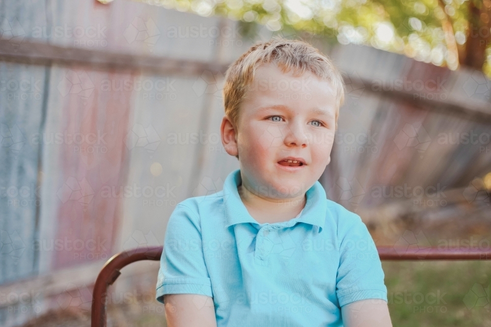 Blond blue eyed 5 year old boy sitting on outdoor bench - Australian Stock Image