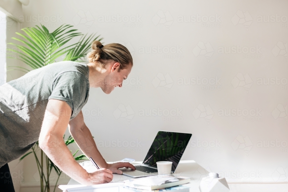 Blond young designer leaning over a desk looking at a laptop screen - Australian Stock Image