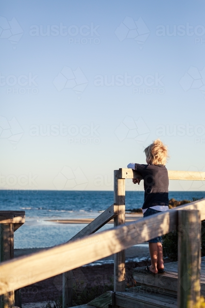blonde boy gazing out over the ocean from wooden stairs to beach - Australian Stock Image