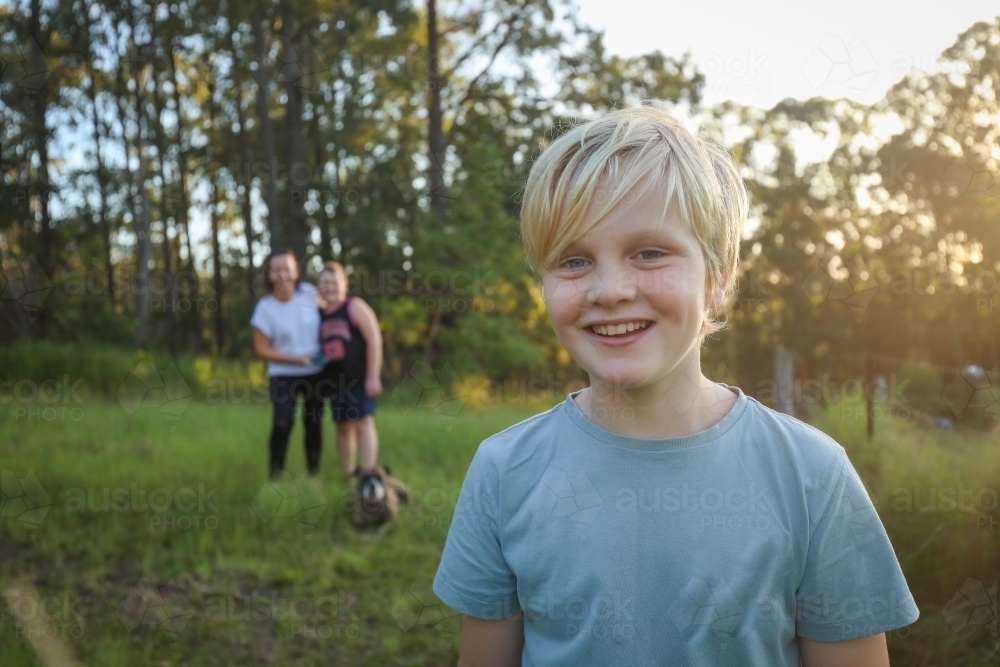 Blonde Caucasian boy smiling in the bush with mother and brother hugging out of focus - Australian Stock Image