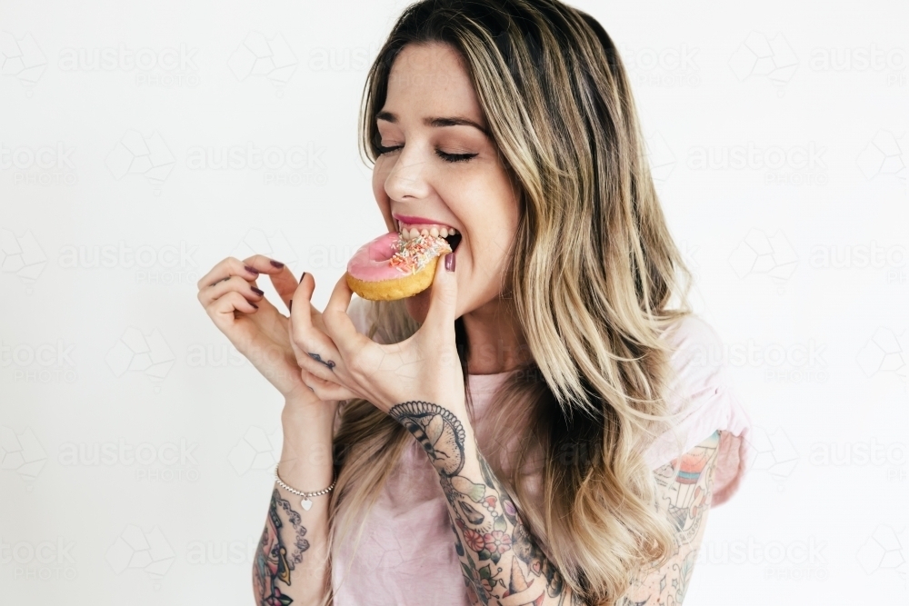 Blonde girl in a pink t shirt biting into a pink iced donut - Australian Stock Image