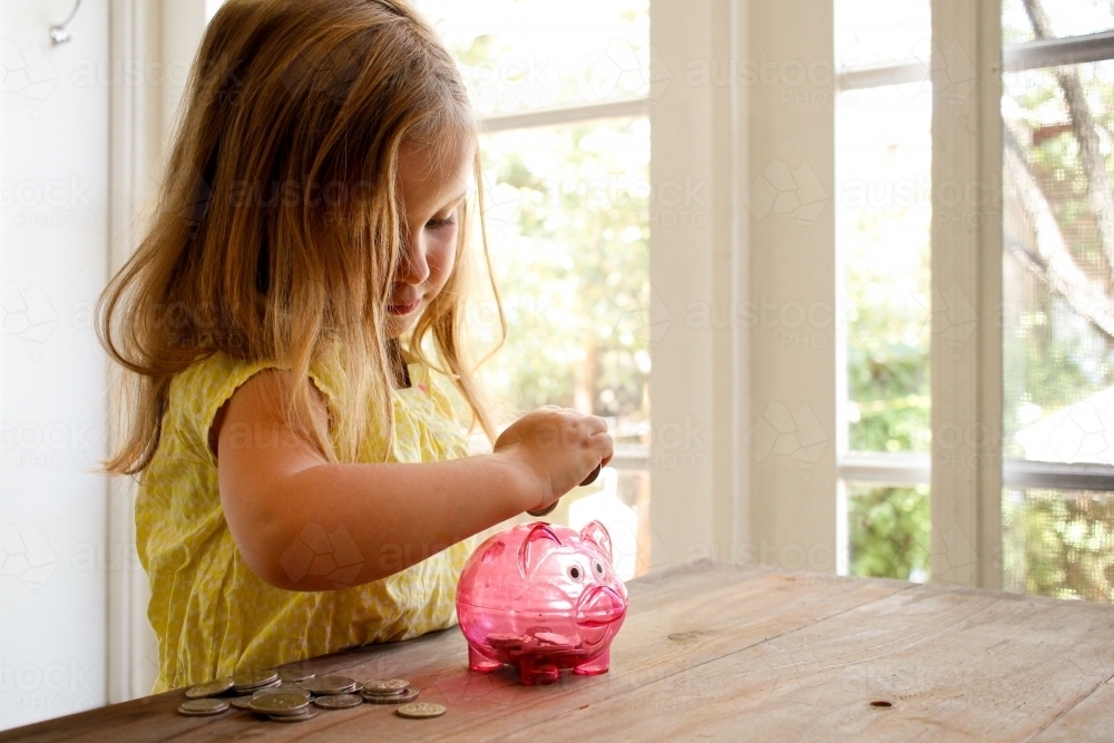 blonde girl wearing yellow blouse putting coins in the pink piggy bank with coins on the table - Australian Stock Image
